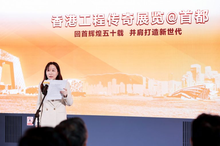 Woman speaking at Hong Kong Engineering Legend Exhibition event with cityscape backdrop.