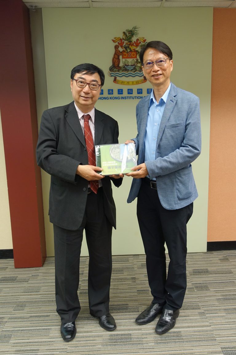 Two men in business attire exchanging a booklet in front of Hong Kong Institution of Engineers signage.