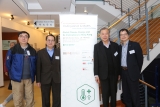Four men standing together at an event, smiling, in front of a white display board with green logo.