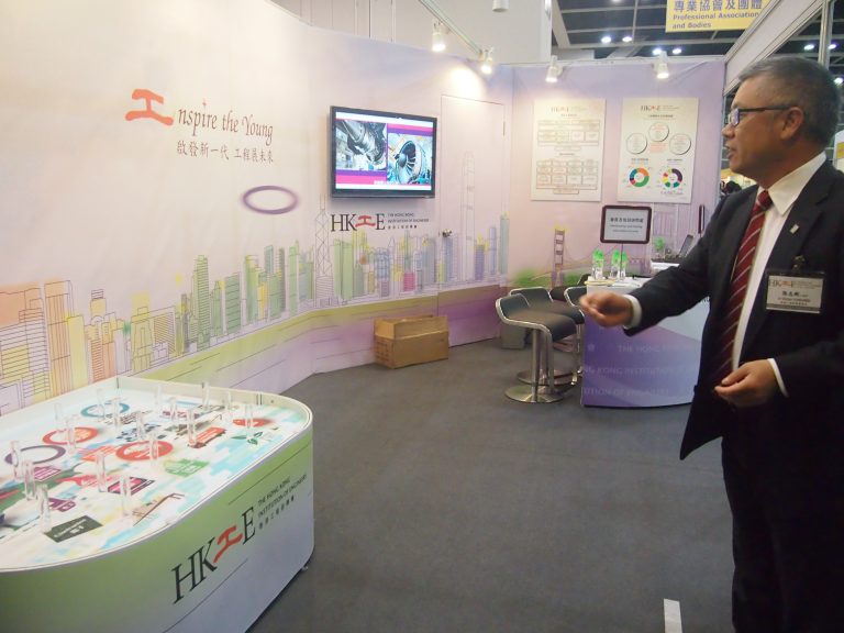 Man gesturing at HKIE exhibition booth with interactive display and informational posters.