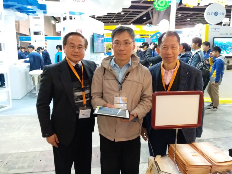 Three men posing at a trade show, one holding a tablet, others wearing event badges.