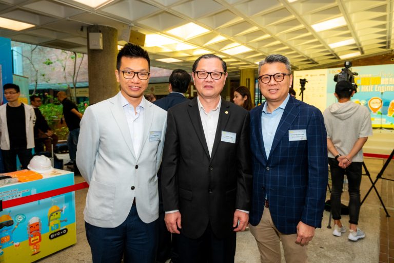 Three men in suits posing together at an event with name tags and a robot display nearby.