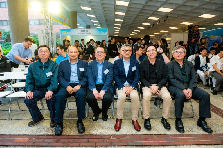 Six men in business attire seated in front of an event crowd.