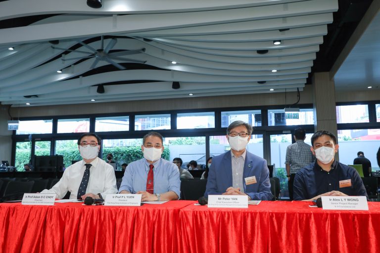 Four men wearing masks seated at a red table with nameplates during an event.