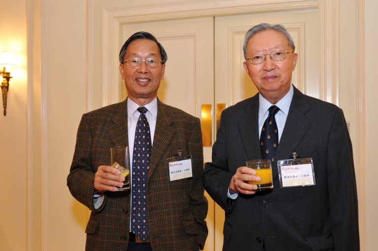Two men in suits holding drinks at an event, wearing name tags.