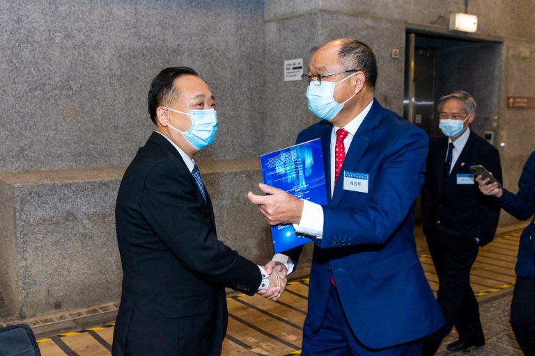 Two men in suits and face masks shake hands while exchanging a blue folder in a hallway.