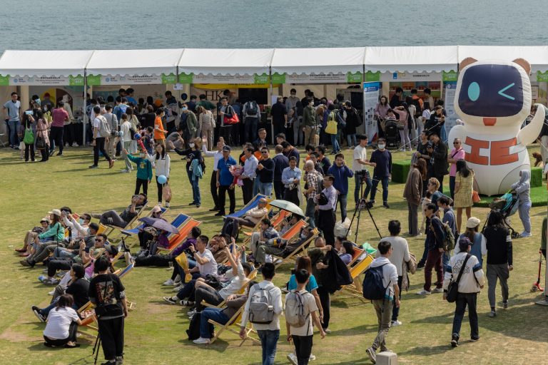 People gather at outdoor event near water, some seated in lounge chairs, others standing near booths and a large inflatable robot.