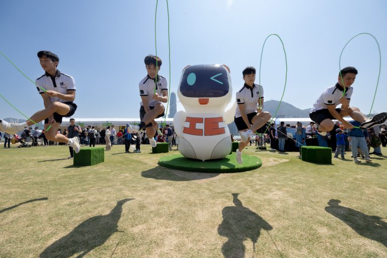 Four athletes jump rope around a large robot mascot on a grassy field.