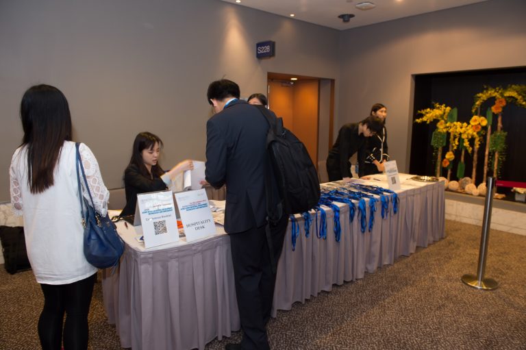 Conference attendees gather at a hospitality desk with informational signs and blue lanyards.