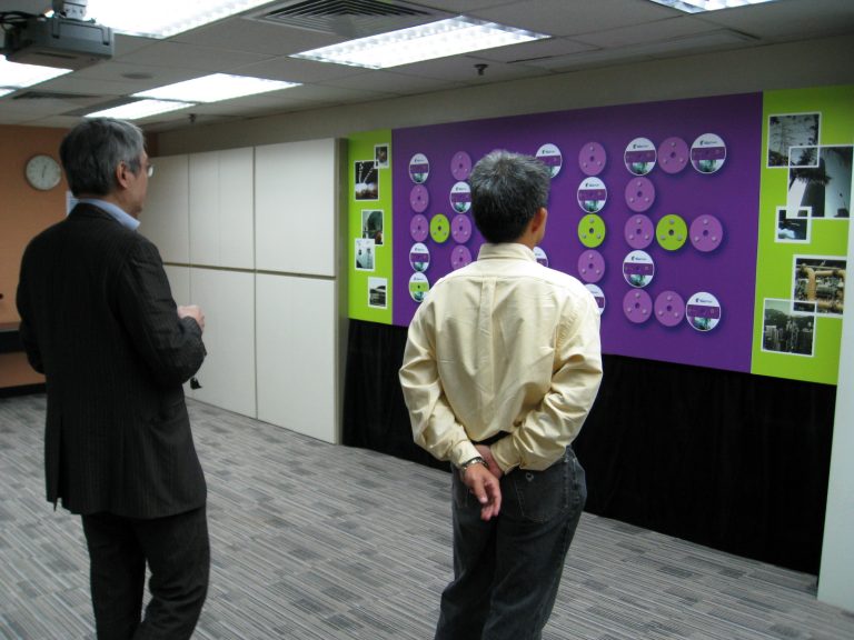 Two men viewing a display wall with circular graphics and photos in an office setting.