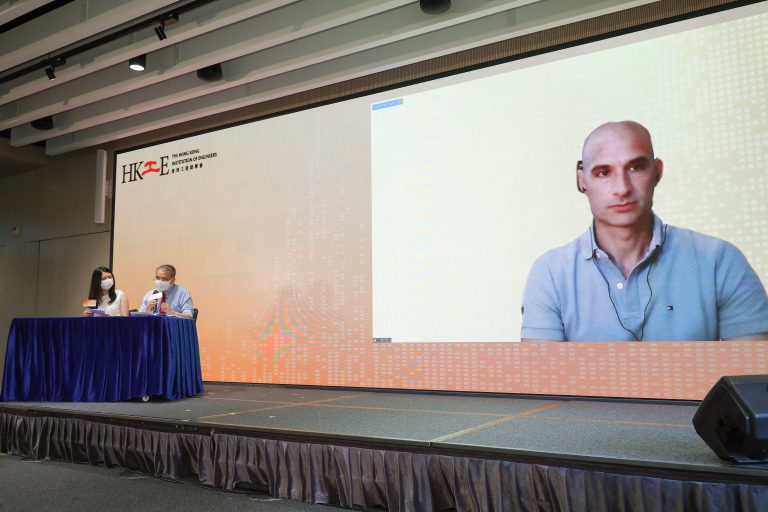 Two speakers at a stage with HKIE logo, large screen shows a man joining remotely.