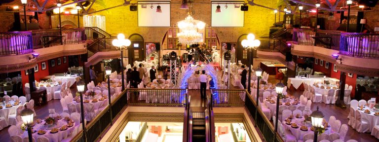 Grand multi-level event hall with tables, chandeliers, and guests at a wedding or banquet.