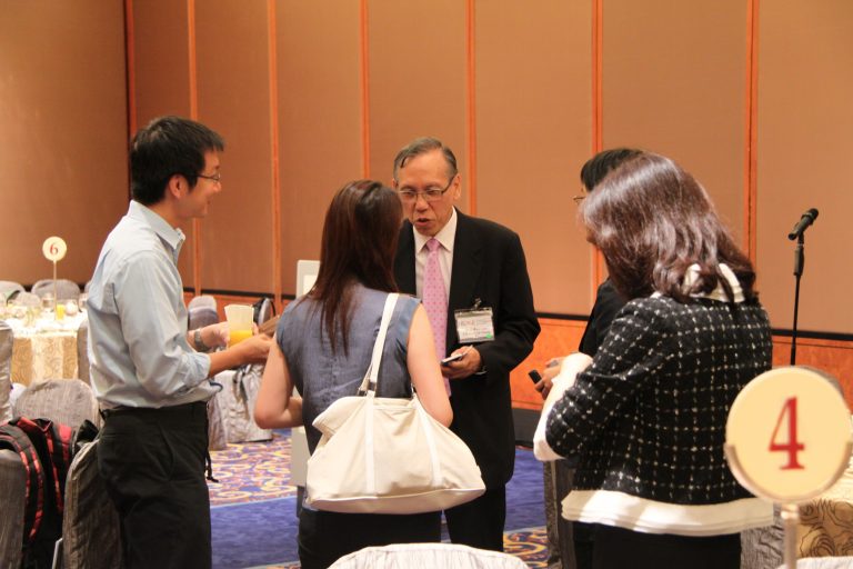 Four people conversing at a conference, one holding a drink, tables with number signs visible.