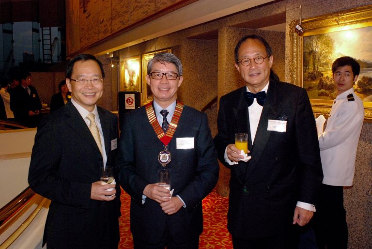 Three men in formal attire posing together at an event, one wearing a ceremonial medal.