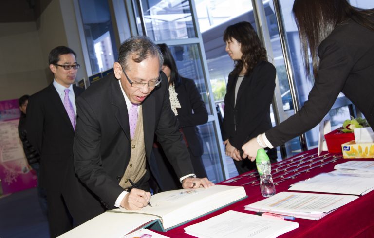 Man signing a guest book at a formal event with colleagues nearby.