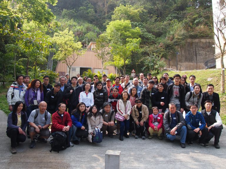 Group of people posing for photo outdoors in a park-like setting with trees and a building in background.