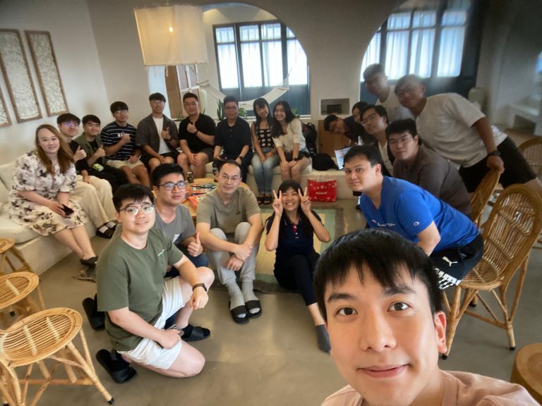 Group of people posing for a selfie in a cozy, well-lit room with wicker furniture.