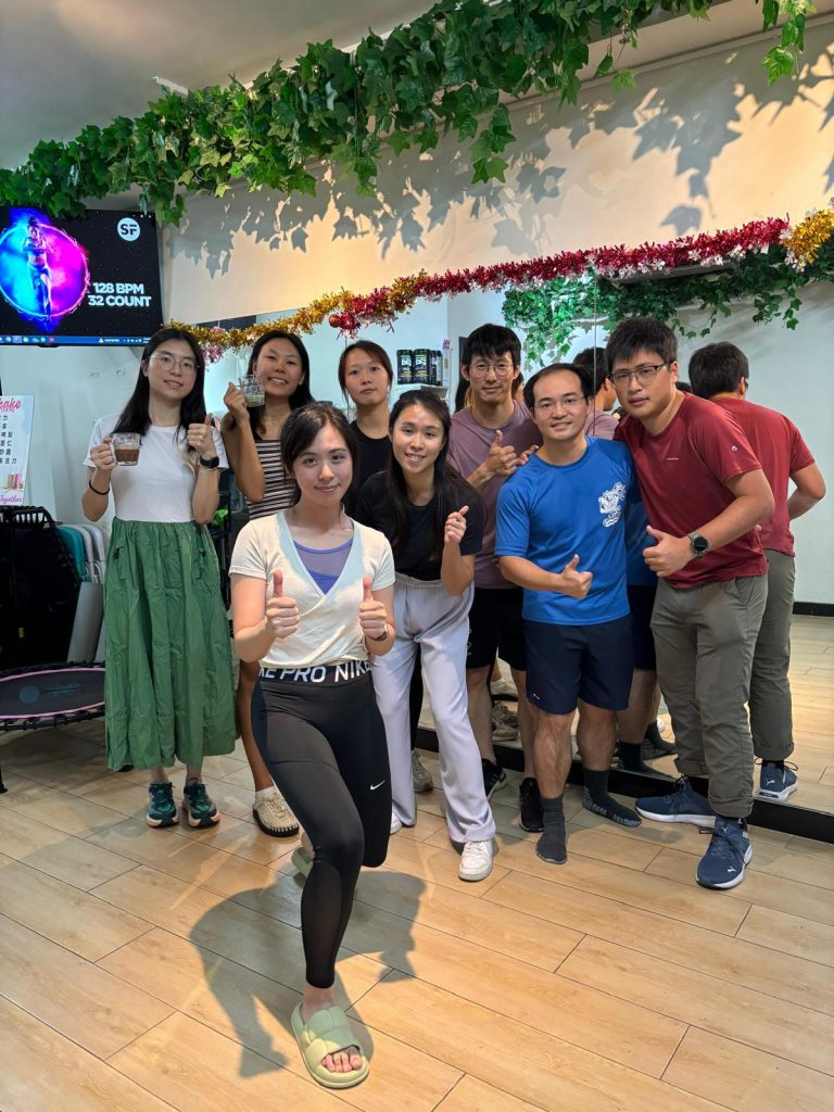Group of people posing with thumbs up in a fitness studio with decorative greenery and a dance-themed screen.