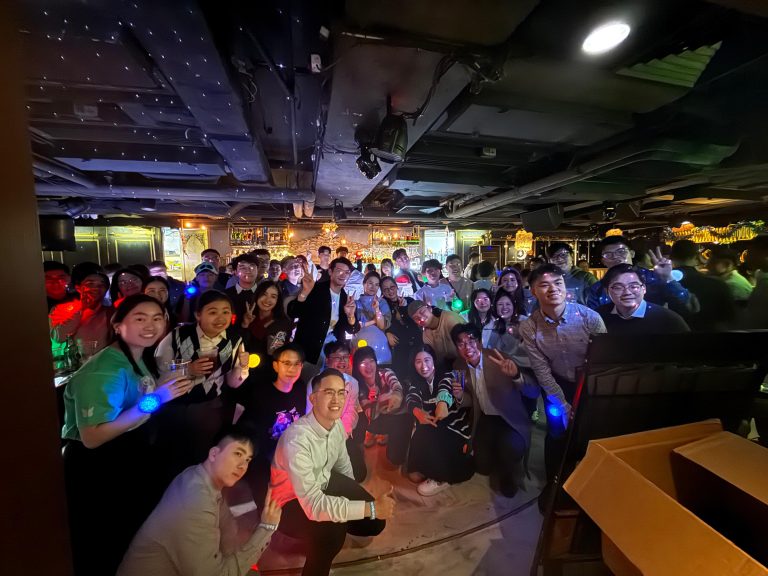 Group of friends posing for photo in dimly lit bar with colorful lights and drinks.