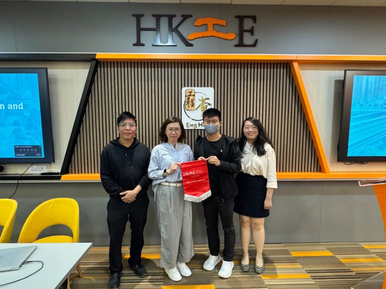 Four people posing for a photo in an office, holding a red HK工E banner.
