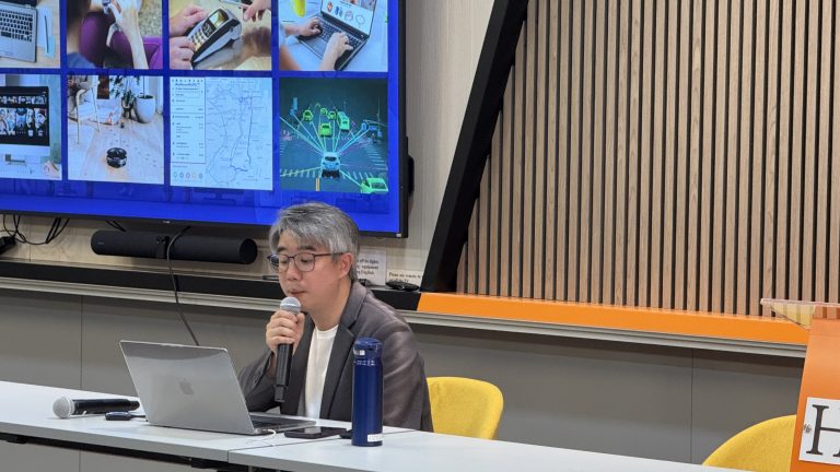 Man speaking into microphone at presentation with large screen displaying tech visuals behind him.