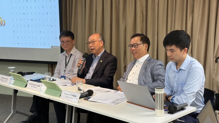 Four men seated at a conference table, one speaking into a microphone.